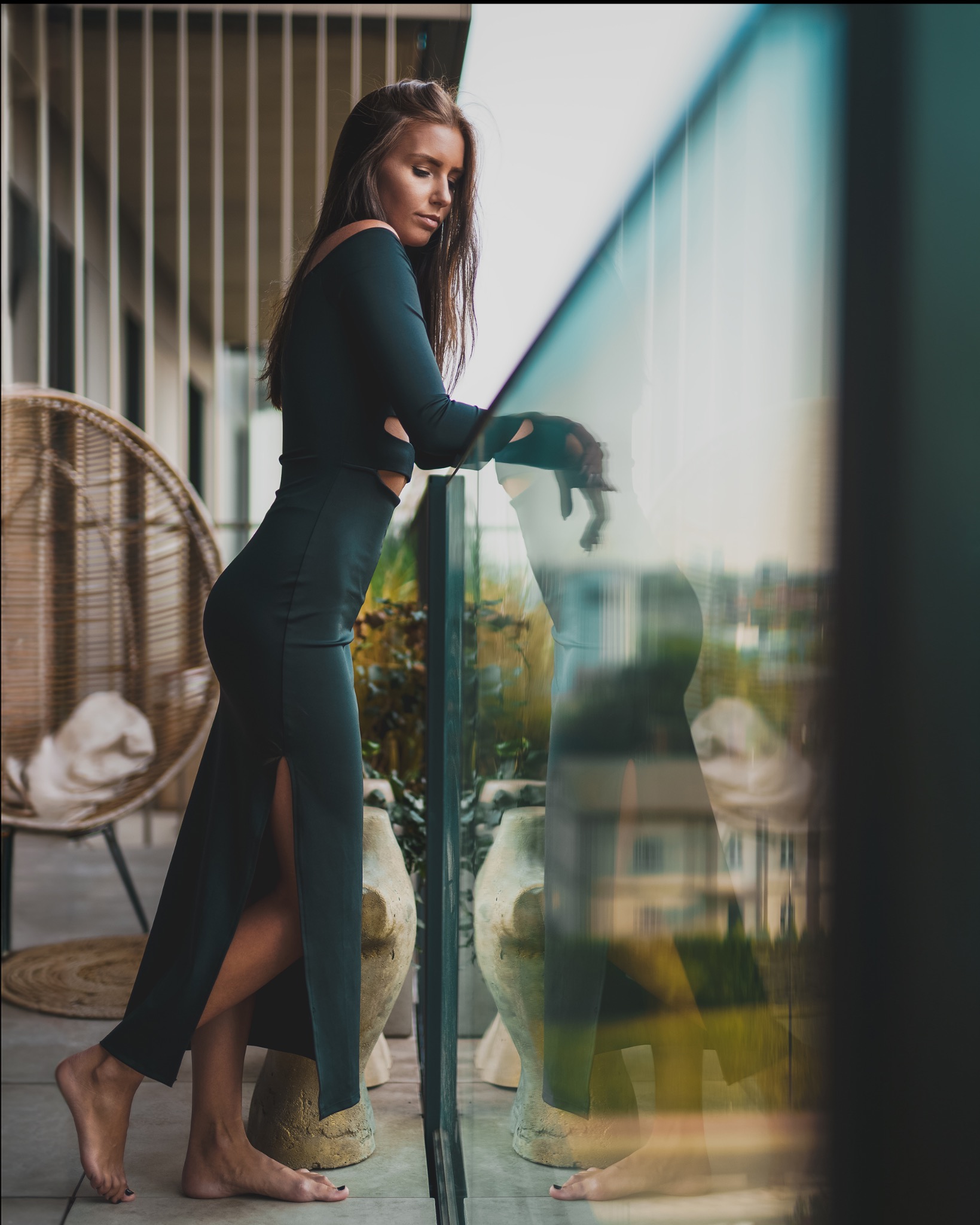 Kobieta w długiej, zielonej sukience z rozcięciem, oparta o szklaną balustradę balkonu z widocznym odbiciem miejskiego krajobrazu, obok wiklinowego fotela i donic z roślinami.