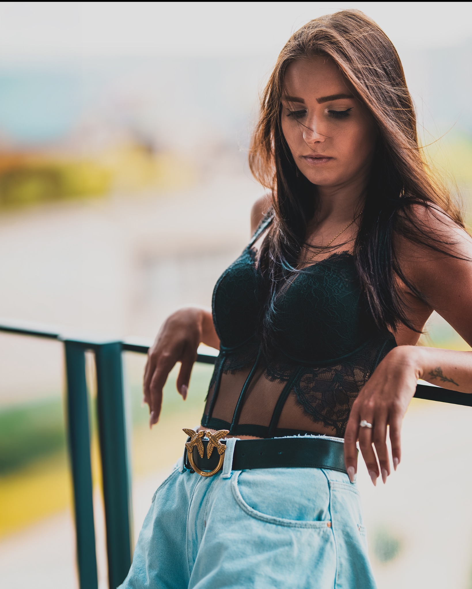 Kobieta w koronkowej bieliźnie i jeansowych szortach opiera się o barierkę, spoglądając w dół, z delikatnym tatuażem na przedramieniu i złotym łańcuszkiem na szyi.