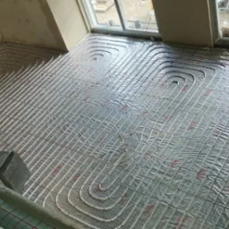 Montaż ogrzewania podłogowego: białe rurki rozprowadzone na srebrnej folii izolacyjnej, widok z góry na poddaszu z oknami dachowymi.