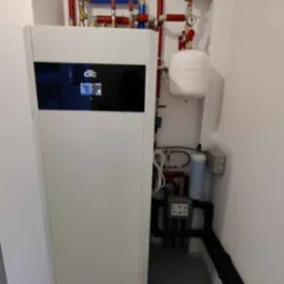 Biała pompa ciepła CTC z instalacją hydrauliczną w tle, widoczne rury w kolorach czerwonym i niebieskim, biały zbiornik wyrównawczy i czarne rury izolowane.