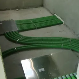 Wiązki zielonych, karbowanych rur instalacyjnych prowadzone pod podłogą, zabezpieczone metalowymi płytami montażowymi w surowym wnętrzu budynku.