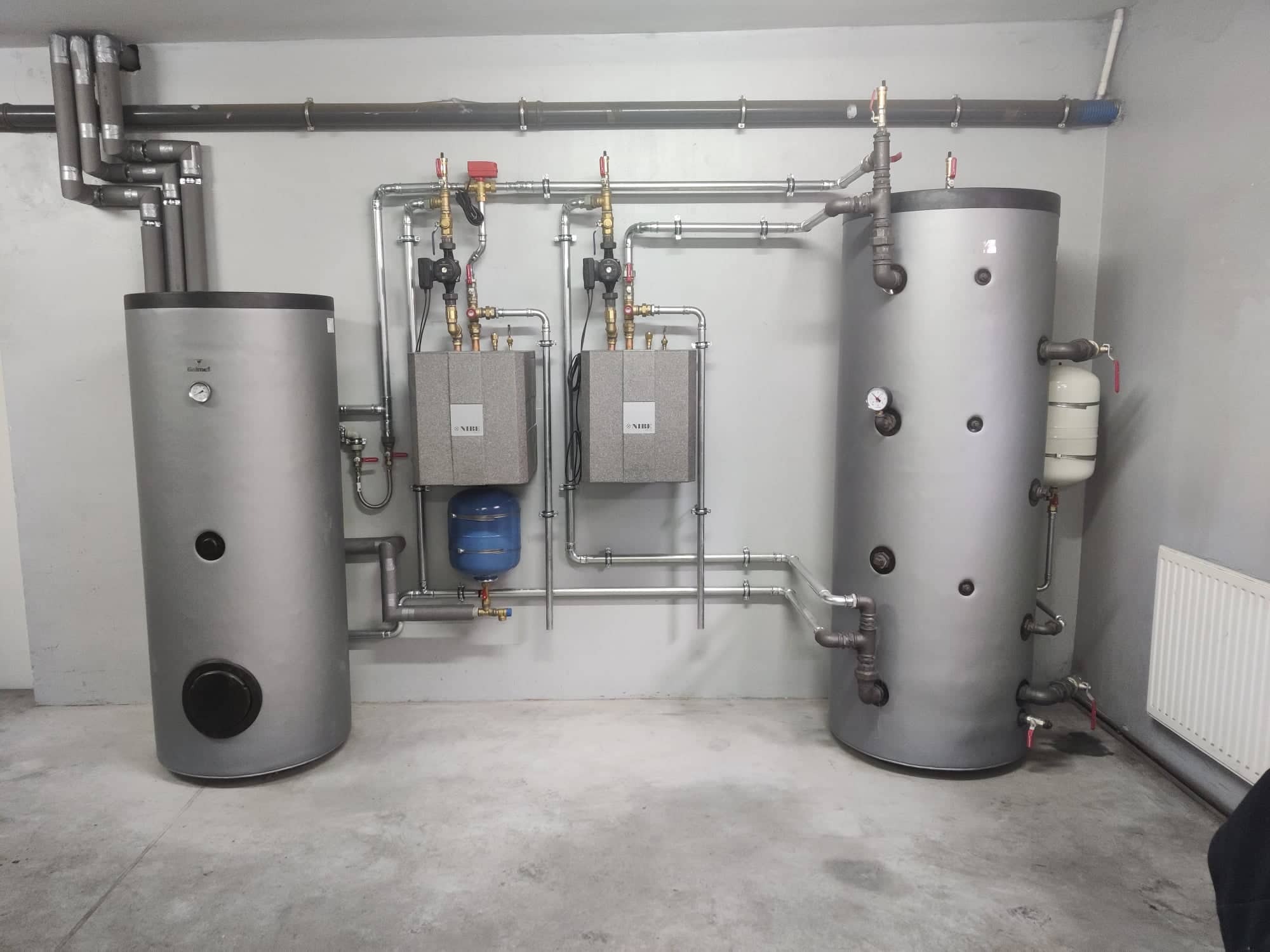 Szara kotłownia z dwoma zbiornikami akumulacyjnymi, dwoma szarymi modułami sterującymi NIBE i niebieskim zbiornikiem wyrównawczym, połączonymi licznymi rurami z zaworami i manometrami.