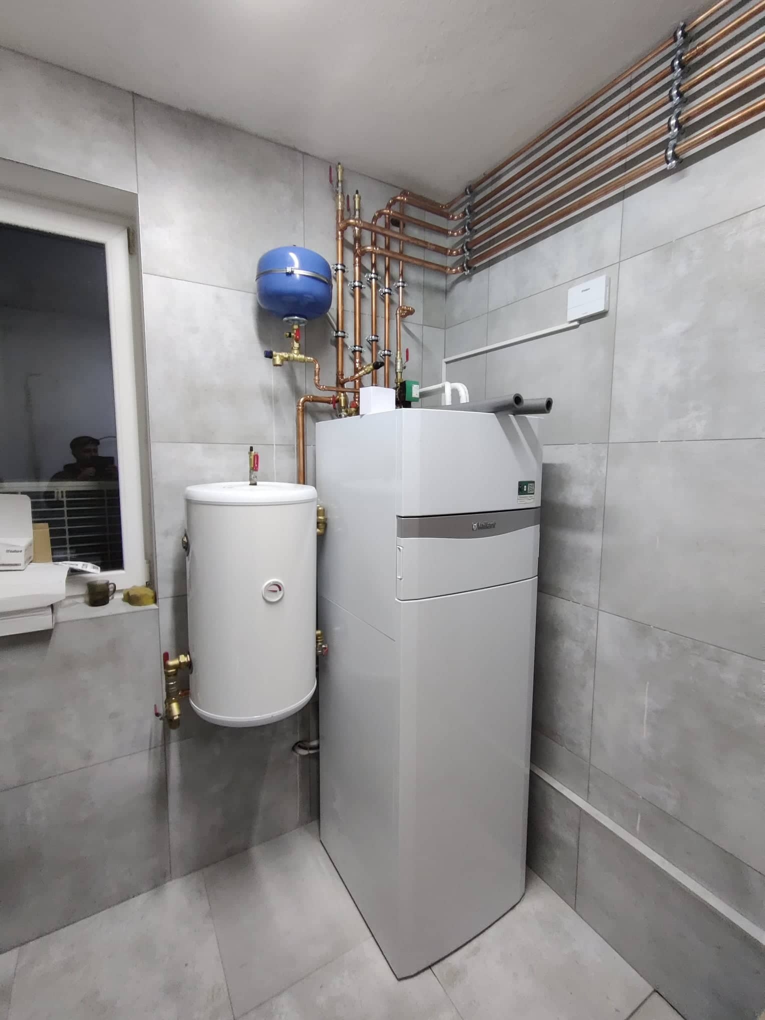 Biały, nowoczesny piec grzewczy Vaillant z zasobnikiem CWU, niebieskim naczyniem przeponowym i miedzianą instalacją rurową na tle szarych płytek ceramicznych w pomieszczeniu z oknem.