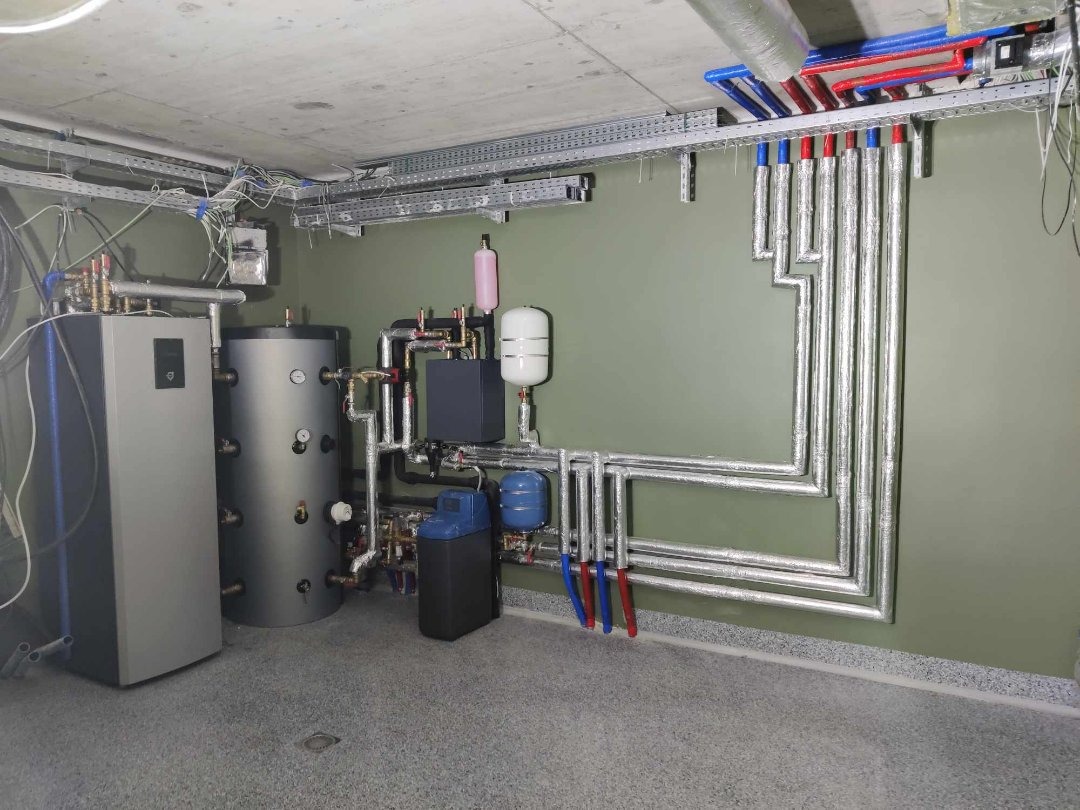 Kotłownia z szarym kotłem, zasobnikiem ciepłej wody i rozbudowaną instalacją rur w izolacji aluminiowej, widoczne także filtry i zbiorniki wyrównawcze.