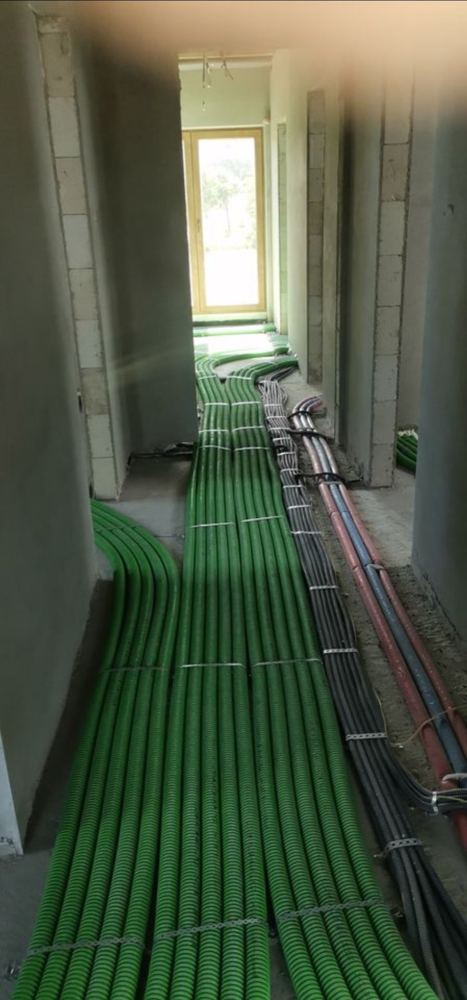 Wiązki zielonych rur karbowanych i kabli elektrycznych ułożone równolegle na podłodze w surowym wnętrzu budynku w trakcie instalacji. Widoczne jasne okno w tle.