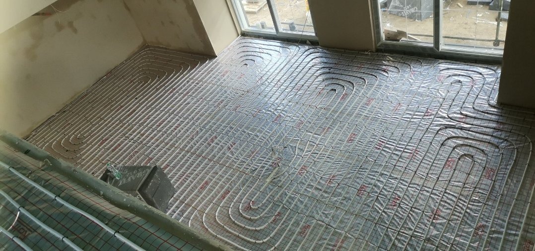 Montaż ogrzewania podłogowego: białe rurki rozprowadzone na srebrnej folii izolacyjnej, widok z góry na poddaszu z oknami dachowymi.