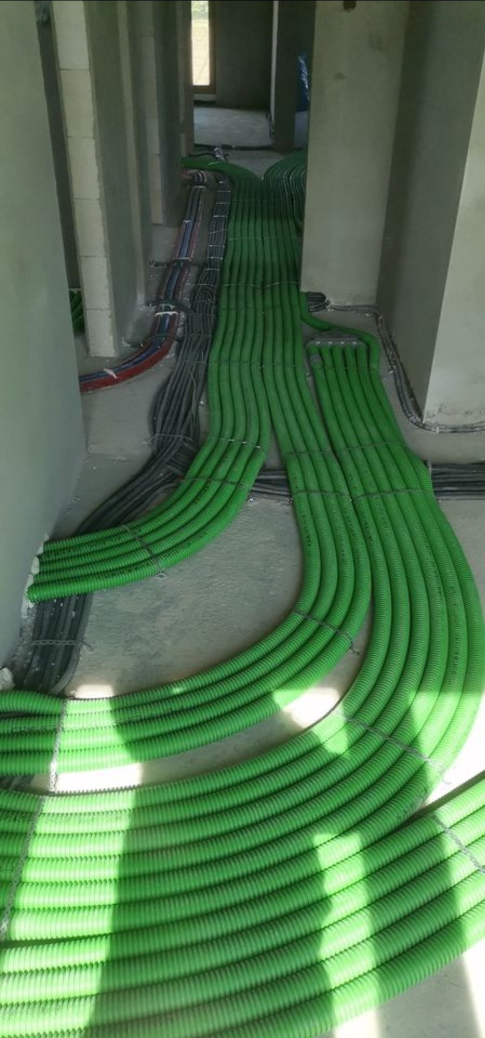 Wnętrze w stanie surowym z ułożonymi rzędami zielonych rur karbowanych na podłodze, obok widoczne czarne i kolorowe przewody, w tle okno.