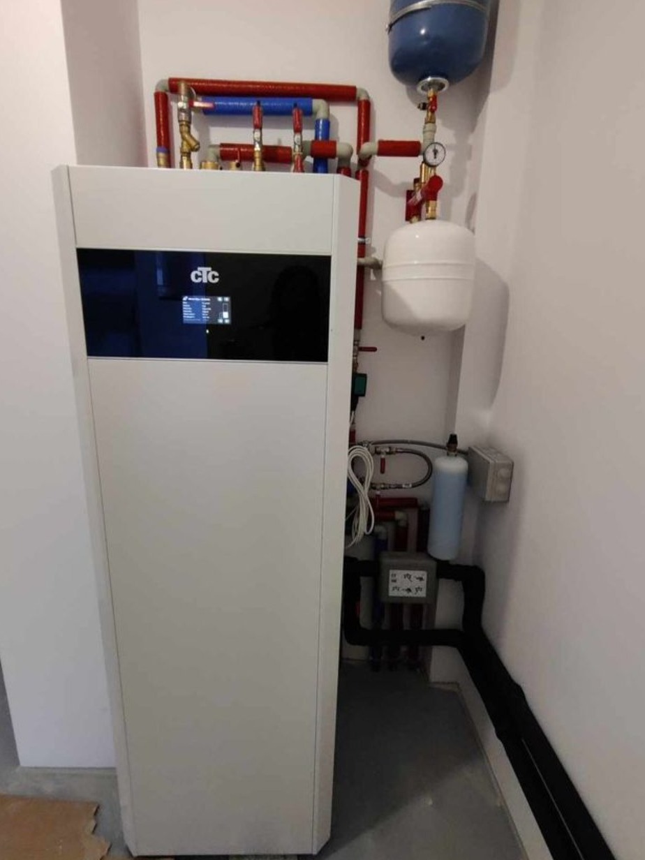 Biała pompa ciepła CTC z instalacją hydrauliczną w tle, widoczne rury w kolorach czerwonym i niebieskim, biały zbiornik wyrównawczy i czarne rury izolowane.