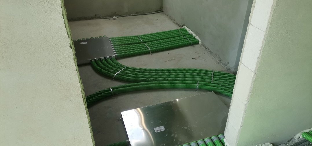Wiązki zielonych, karbowanych rur instalacyjnych prowadzone pod podłogą, zabezpieczone metalowymi płytami montażowymi w surowym wnętrzu budynku.