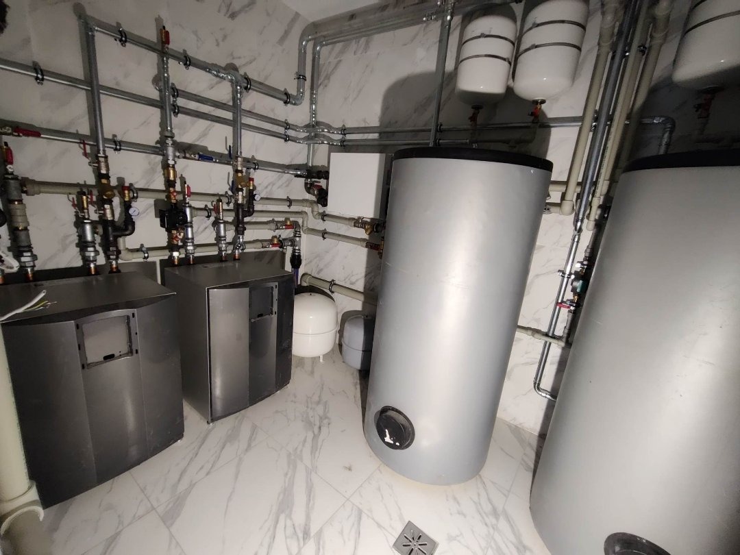 Kotłownia z dwoma zbiornikami akumulacyjnymi, dwoma kotłami i rozbudowaną instalacją rurową z zaworami na marmurowej podłodze i tle marmurowej ściany.