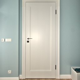 SCHODY I DRZWI ŻĄDŁO SPÓŁKA JAWNA - Białe drzwi wewnętrzne z prostokątnym panelem, chromowaną klamką i czarnym zamkiem, na tle błękitnej ściany z białymi listwami przypodłogowymi i drewnianą podłogą.