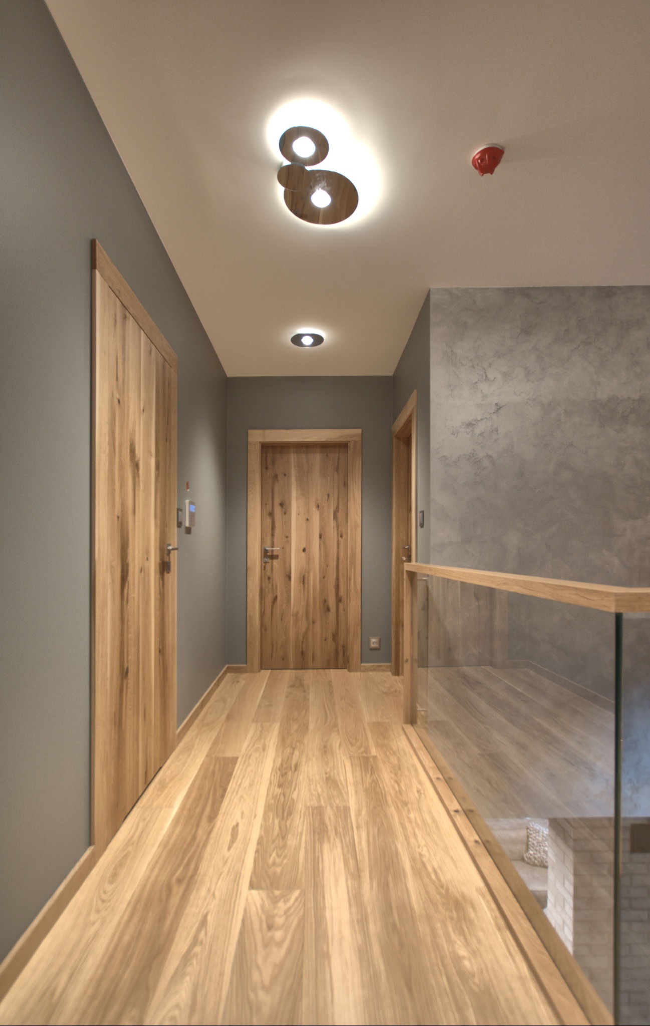 Korytarz z dębowymi drzwiami i podłogą, szare ściany, nowoczesne oświetlenie sufitowe, szklana balustrada z drewnianą poręczą.