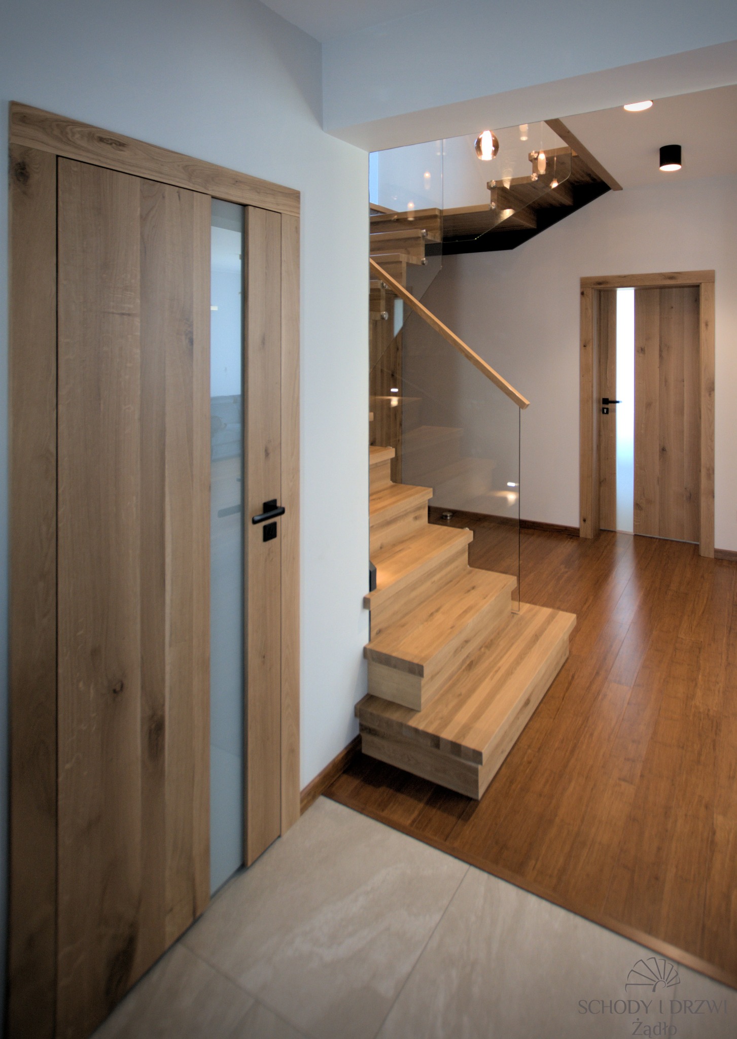 Nowoczesne wnętrze z drewnianymi schodami z balustradą szklaną i dwiema parami drzwi drewnianych z matowym przeszkleniem oraz ciemnymi klamkami, widoczne oświetlenie punktowe.