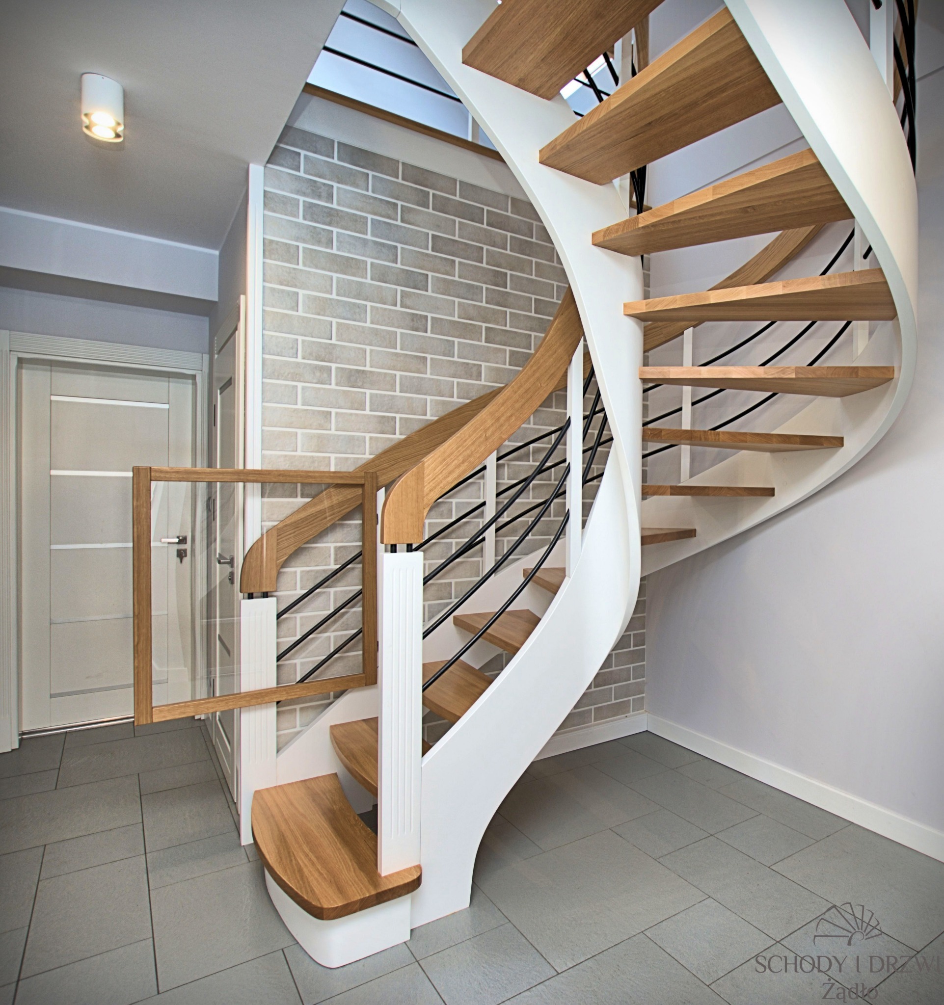 Spiralne schody z jasnego drewna i białymi elementami konstrukcyjnymi, zabezpieczone czarnymi, metalowymi prętami, z dodatkowym zabezpieczeniem w postaci drewnianej barierki z przezroczystym...