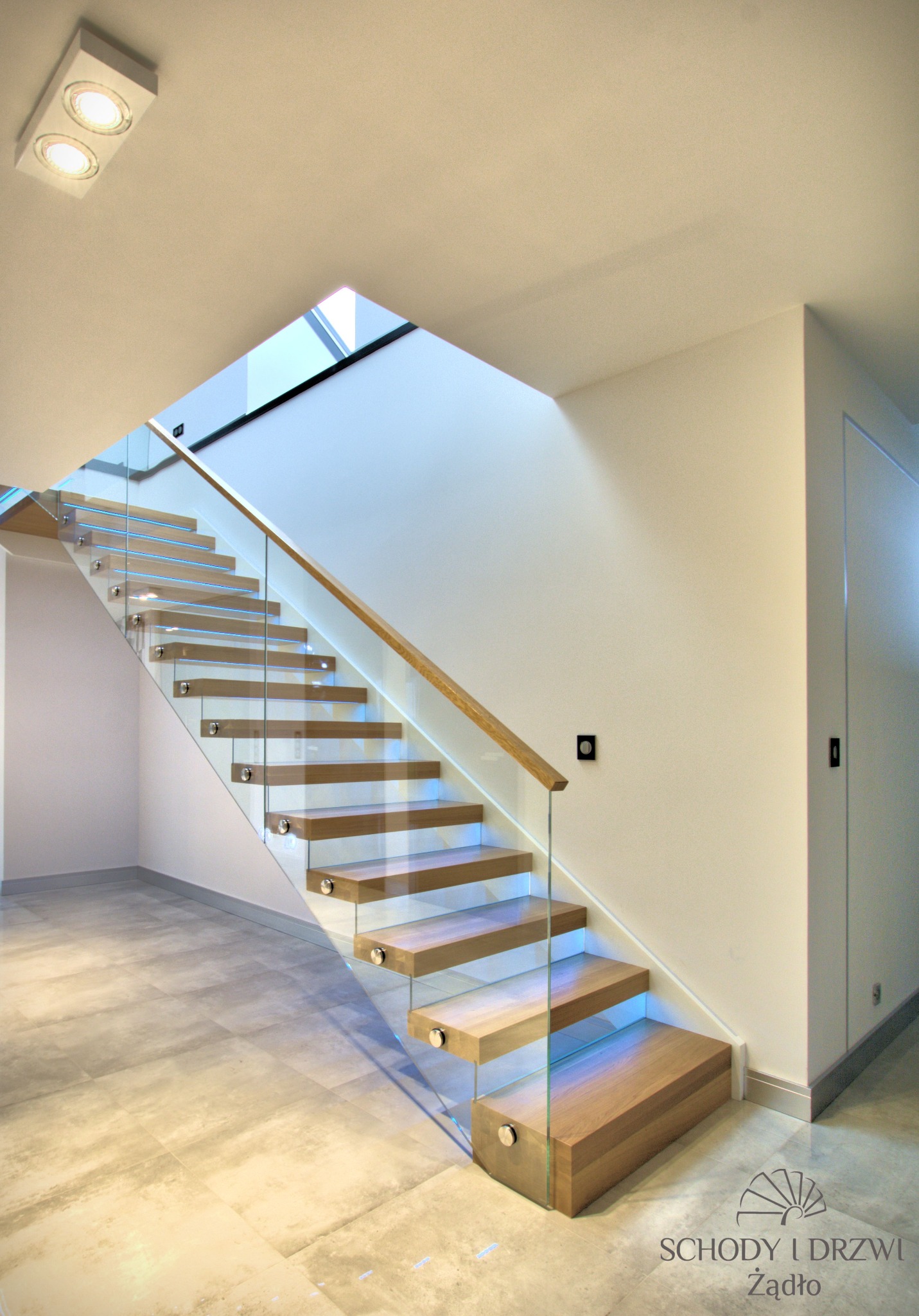 Nowoczesne schody z drewnianymi stopniami i szklaną balustradą, mocowane na metalowych elementach, w jasnym wnętrzu z szarymi płytkami podłogowymi i subtelnym oświetleniem.