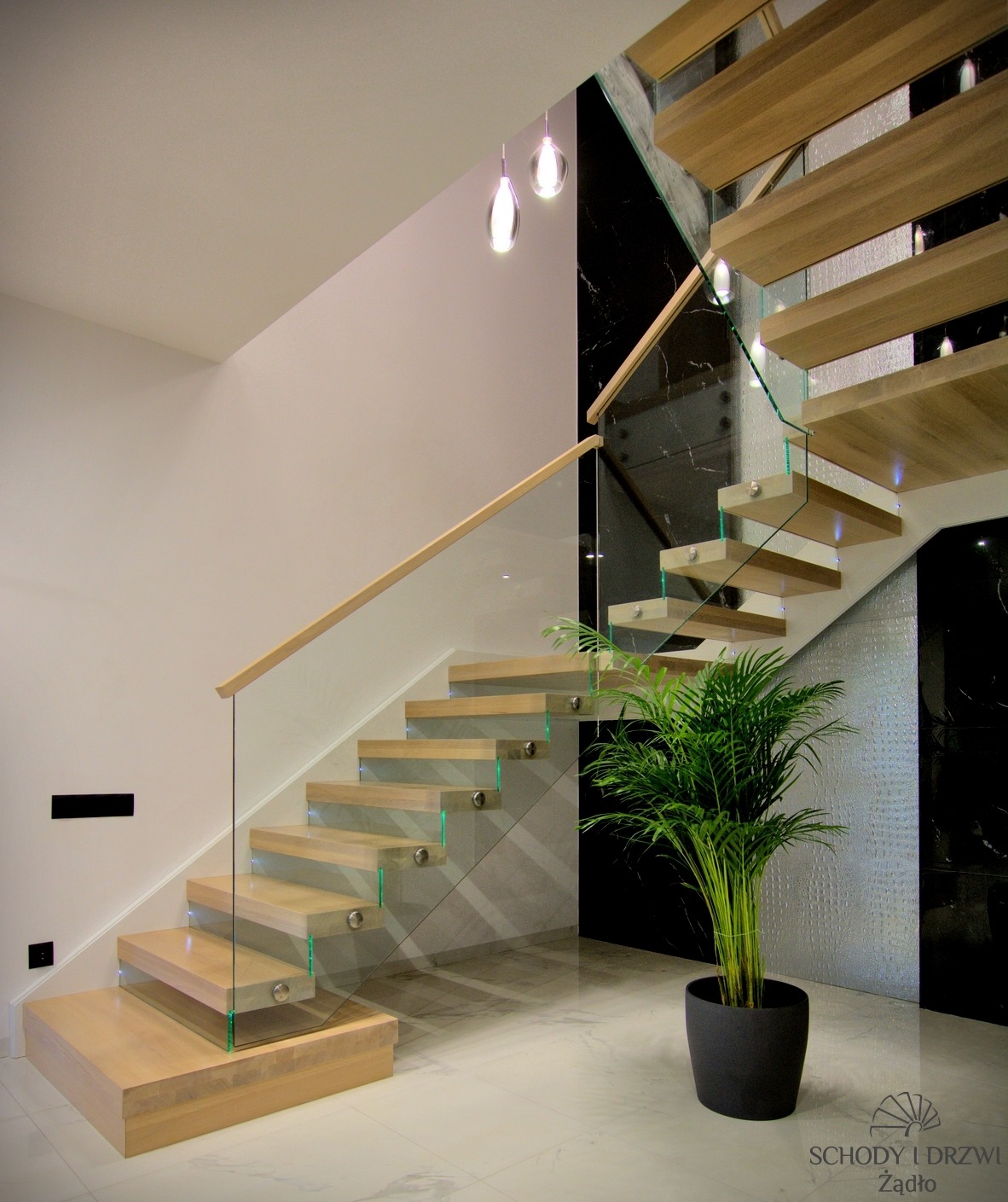 Nowoczesne schody z jasnego drewna z przezroczystymi szklanymi balustradami i drewnianą poręczą, oświetlone wiszącymi lampami, z rośliną doniczkową u podnóża, na tle marmurowej ściany.
