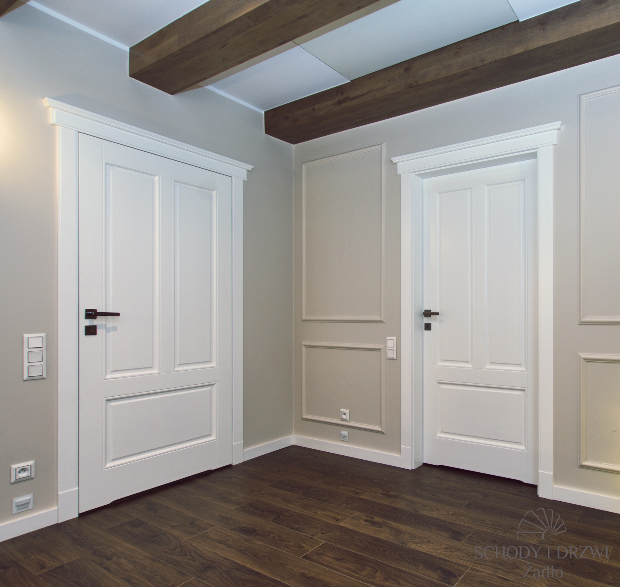 Dwa białe, panelowe drzwi wewnętrzne z czarnymi klamkami, widoczne w rogu pokoju z drewnianą podłogą i dekoracyjnymi listwami ściennymi. Drewniane belki stropowe dodają charakteru wnętrzu.