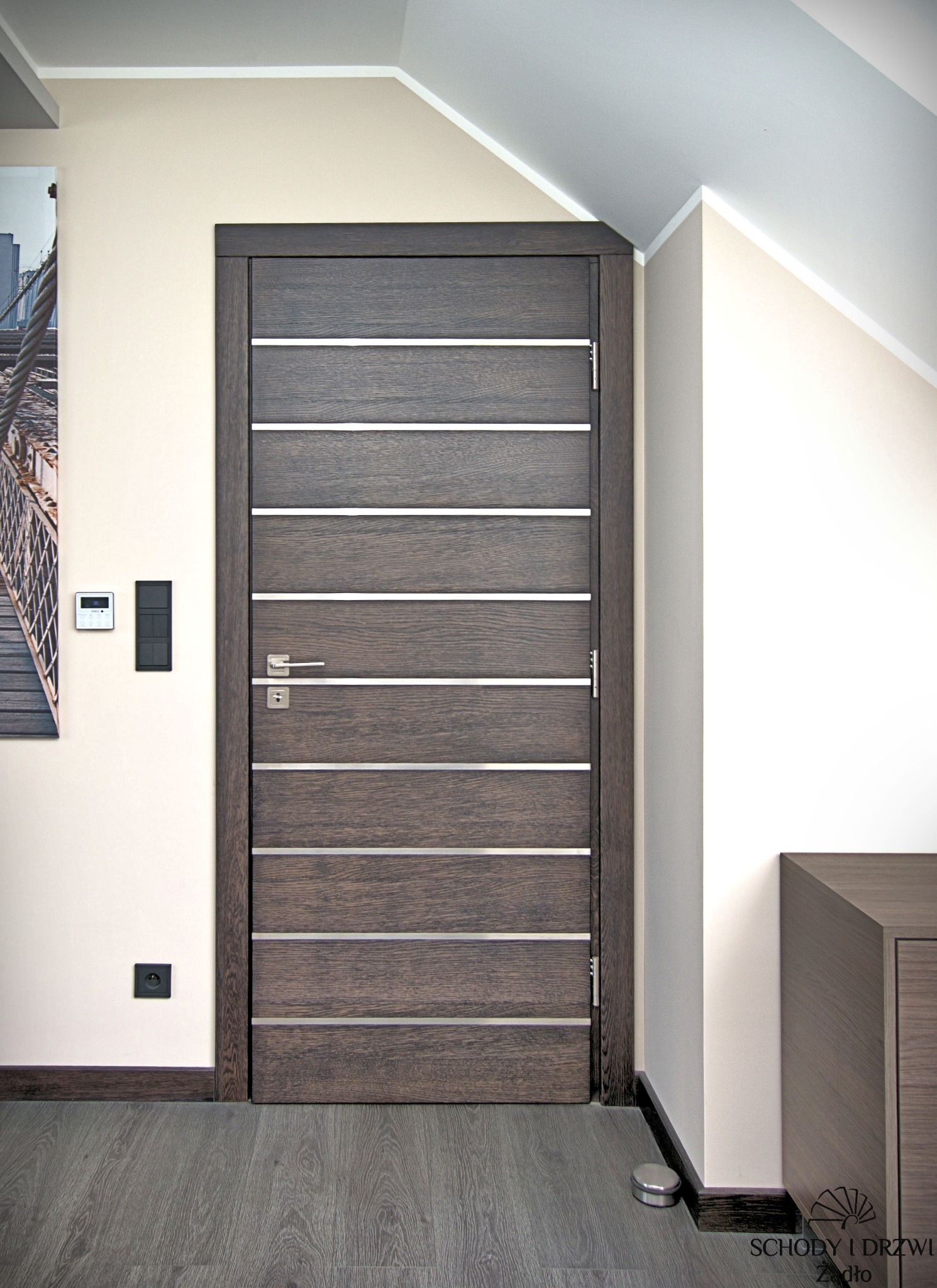 Ciemne drzwi drewniane z poziomymi, srebrnymi wstawkami, zamontowane w jasnym wnętrzu z poddaszem, widoczny fragment obrazu na ścianie i czarne włączniki.