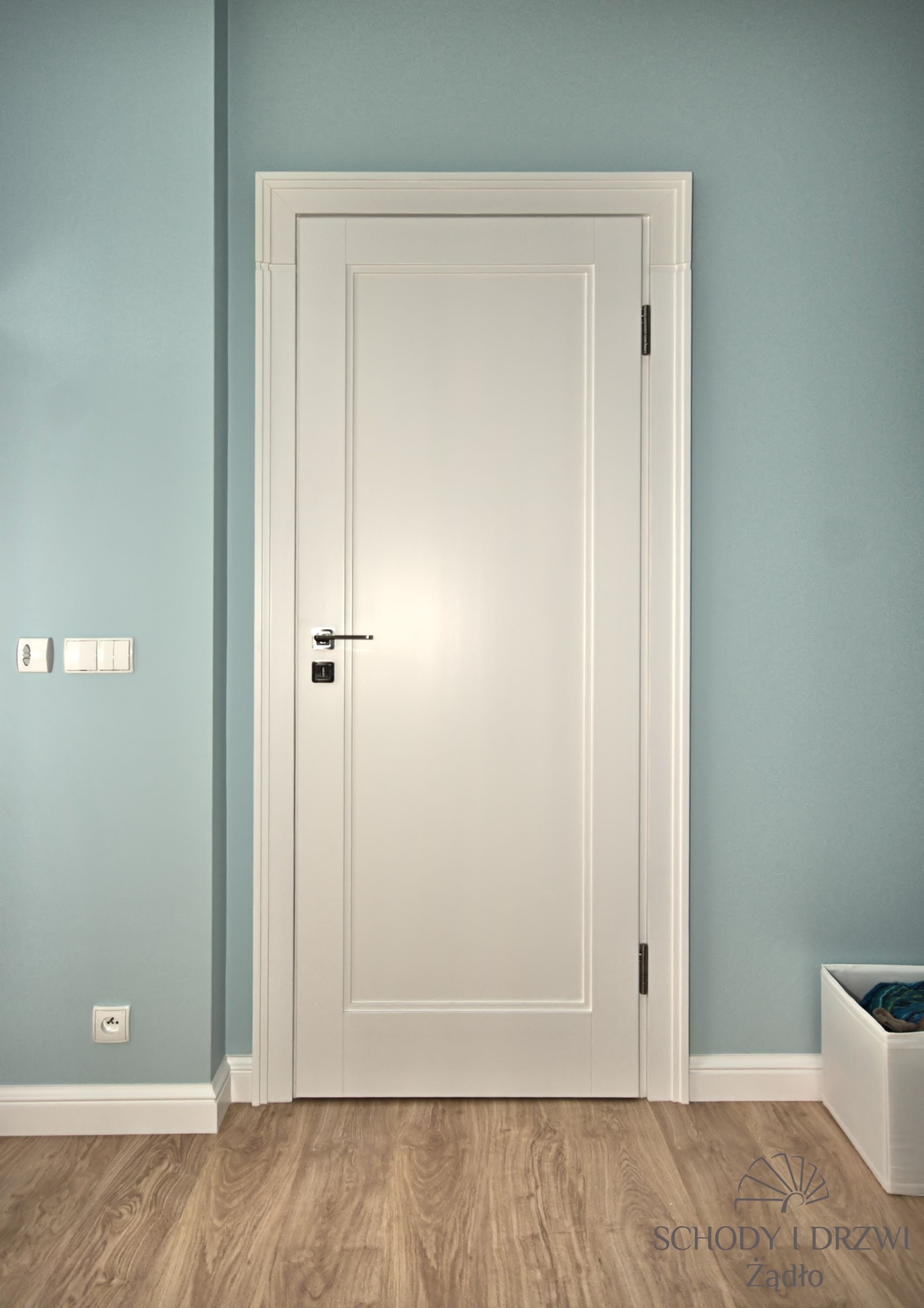 Białe drzwi wewnętrzne z prostokątnym panelem, chromowaną klamką i czarnym zamkiem, na tle błękitnej ściany z białymi listwami przypodłogowymi i drewnianą podłogą.