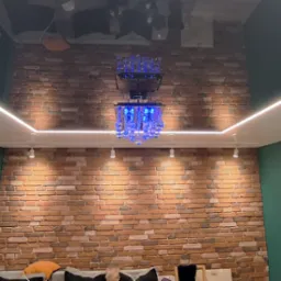 Pokój z ceglaną ścianą, oświetleniem LED pod sufitem i dekoracyjnym żyrandolem z niebieskimi elementami, odbijającym się w błyszczącym suficie.