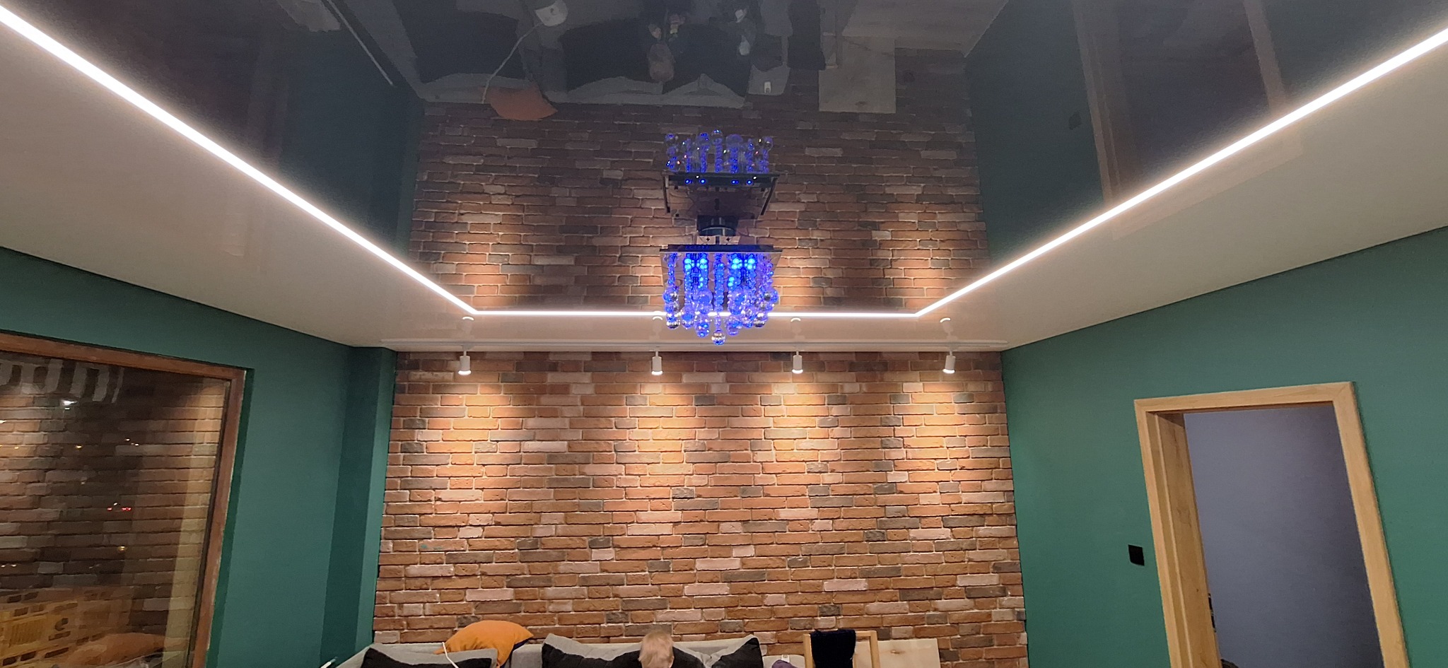 Pokój z ceglaną ścianą, oświetleniem LED pod sufitem i dekoracyjnym żyrandolem z niebieskimi elementami, odbijającym się w błyszczącym suficie.