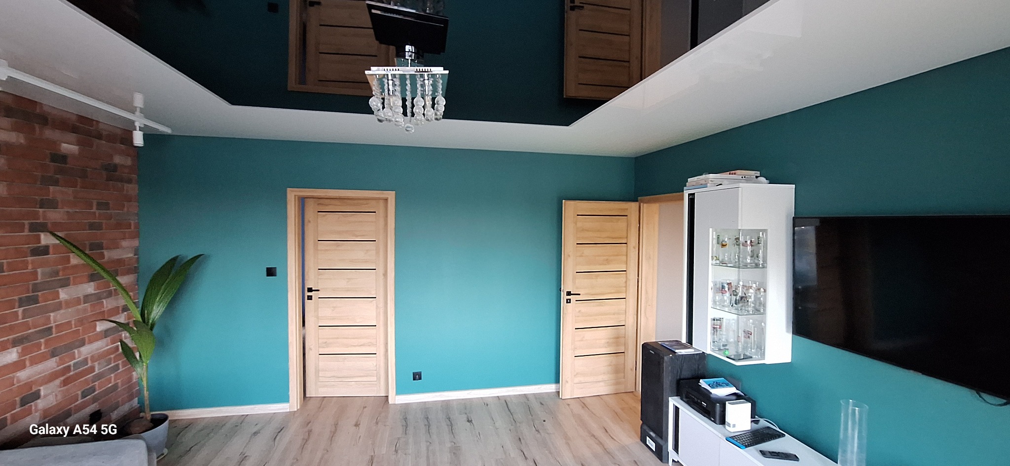 Turkusowa ściana w salonie z dwiema jasnymi drewnianymi drzwiami z poziomymi wstawkami, parkiet, ceglana ściana z rośliną i ozdobny żyrandol.