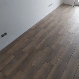 Remont mieszkań - Ułożona podłoga z płytek imitujących drewno w nowym pomieszczeniu, widoczne gniazdka elektryczne na ścianie i fragment okna.