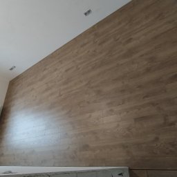 Remont mieszkań - Ściana wykończona panelami imitującymi drewno w jasnym odcieniu, widoczne punkty oświetleniowe na suficie i okno z widokiem na budynki.