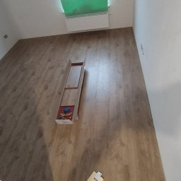 Remont mieszkań - Układanie paneli podłogowych w pokoju, widoczny fragment ułożonej podłogi, otwarte opakowanie z panelami i narzędziami do montażu przy grzejniku.