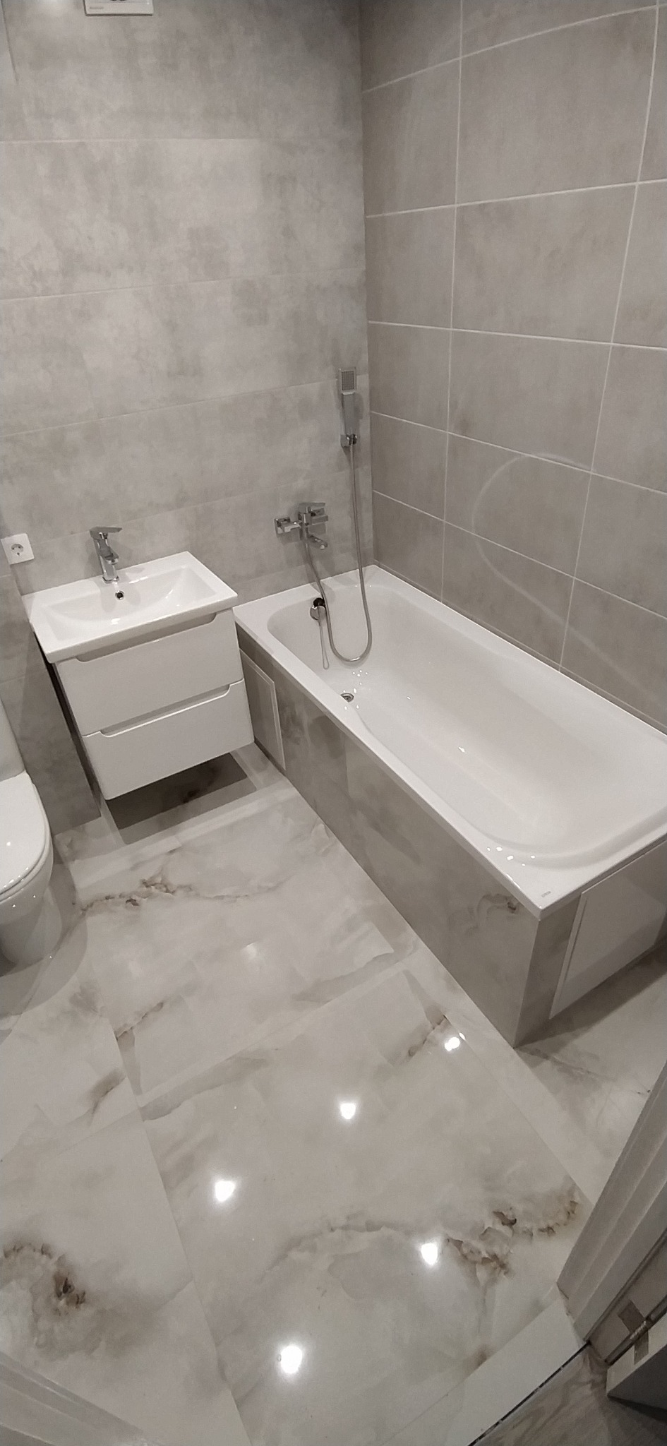 Wykończona łazienka w szarościach: płytki imitujące kamień na ścianach i podłodze, biała wanna obudowana płytkami, biała szafka z umywalką i toaleta.