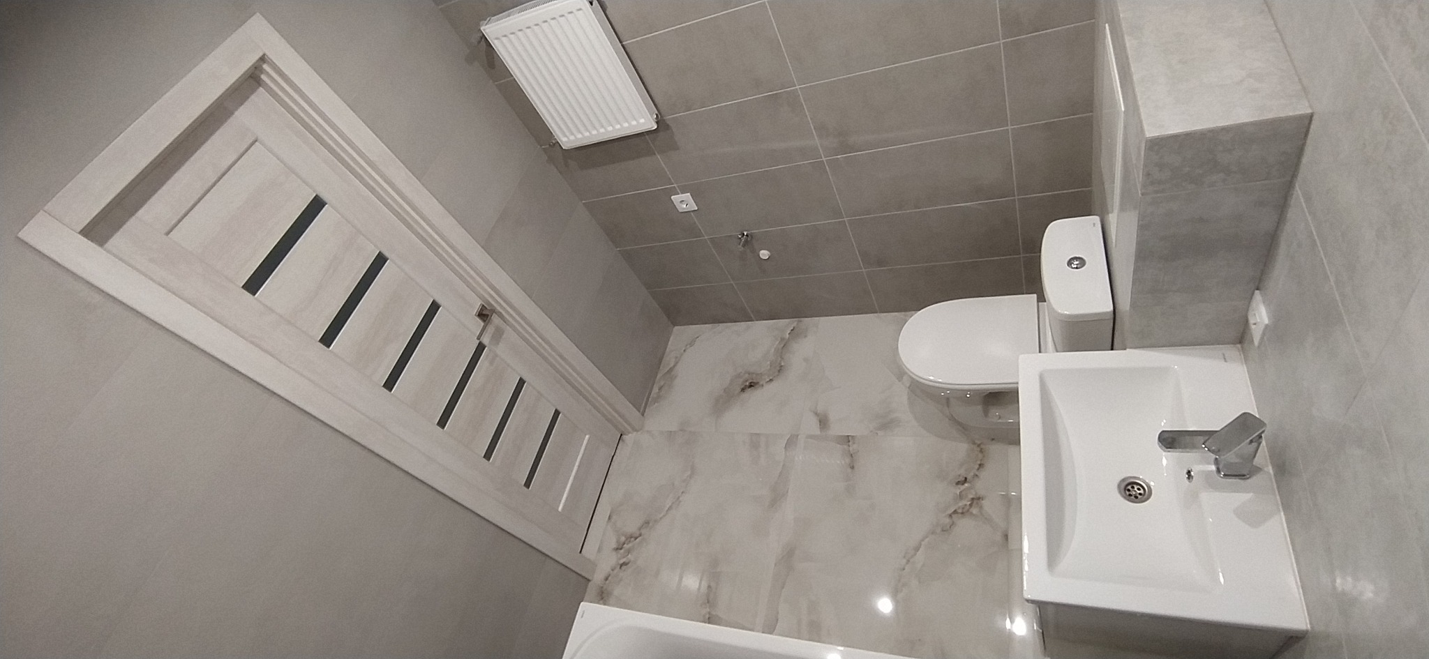 Nowoczesna łazienka w szarościach z białą armaturą, płytkami imitującymi marmur na podłodze i drewnianymi drzwiami z czarnymi wstawkami.