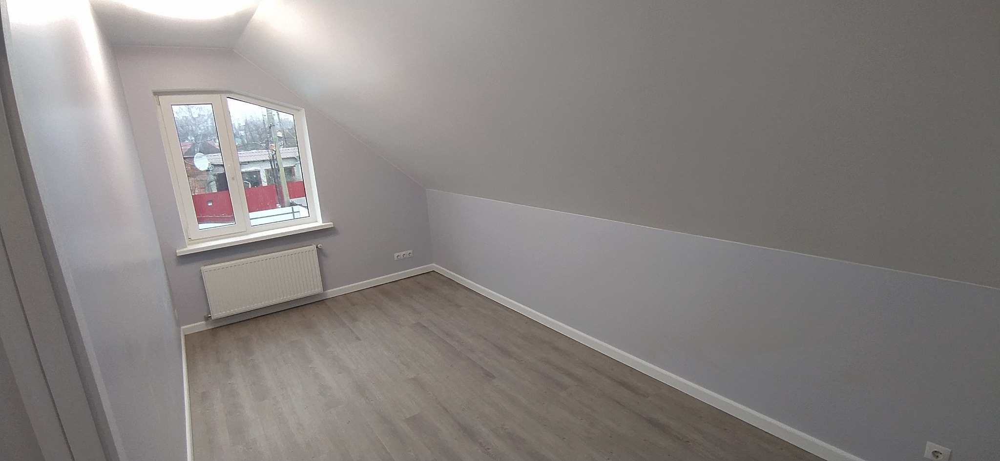 Poddasze po remoncie w odcieniach szarości: gładkie ściany, jasna podłoga imitująca drewno, białe okno dachowe z widokiem na zewnątrz i grzejnik pod nim.