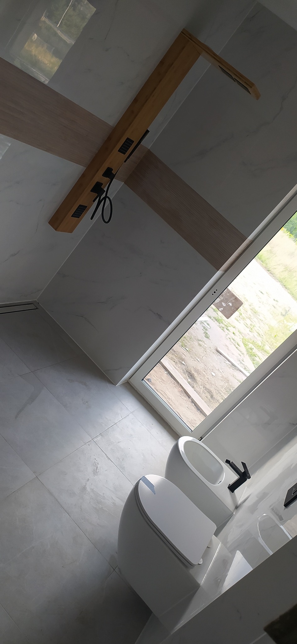 Nowoczesna łazienka z białymi płytkami imitującymi marmur, wiszącą toaletą i bidetem z czarną armaturą, drewnianą belką sufitową z czarnym deszczownicą i oknem z widokiem na zewnątrz.