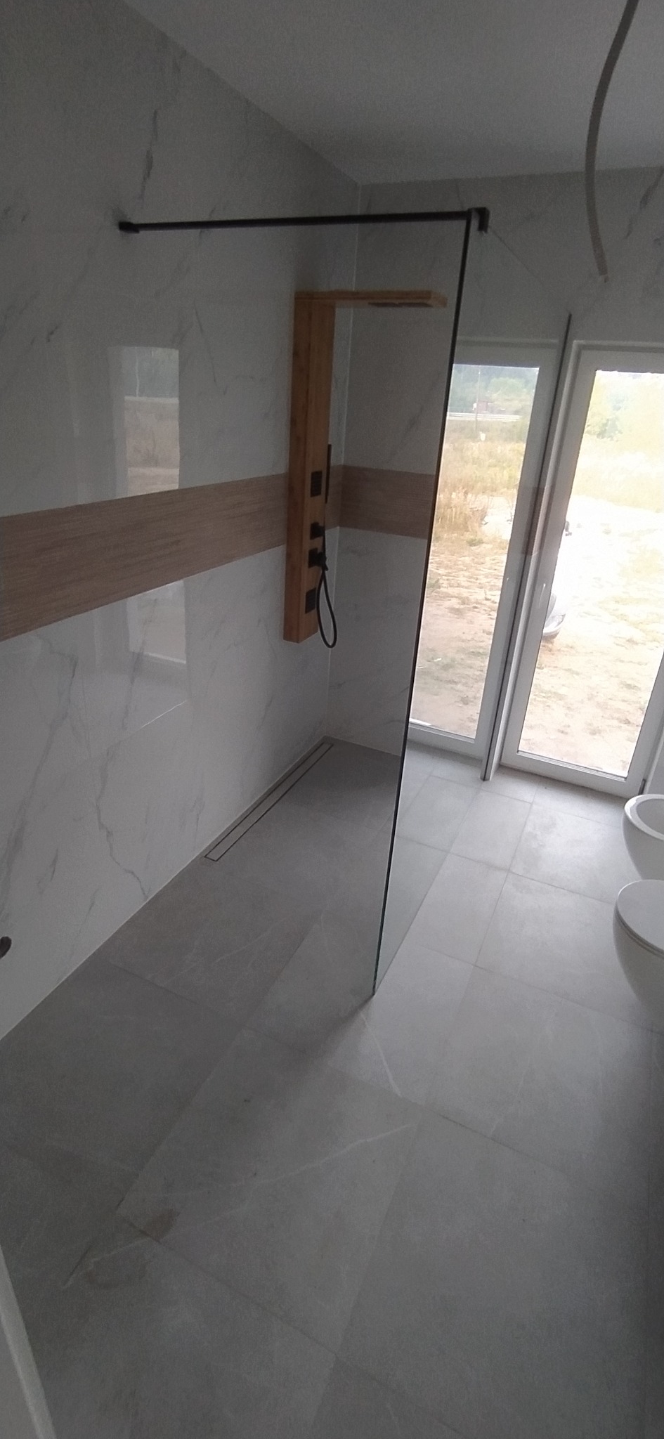 Nowoczesna łazienka z kabiną prysznicową typu walk-in, drewnianym panelem z hydromasażem, białymi płytkami imitującymi marmur i szarymi płytkami podłogowymi.