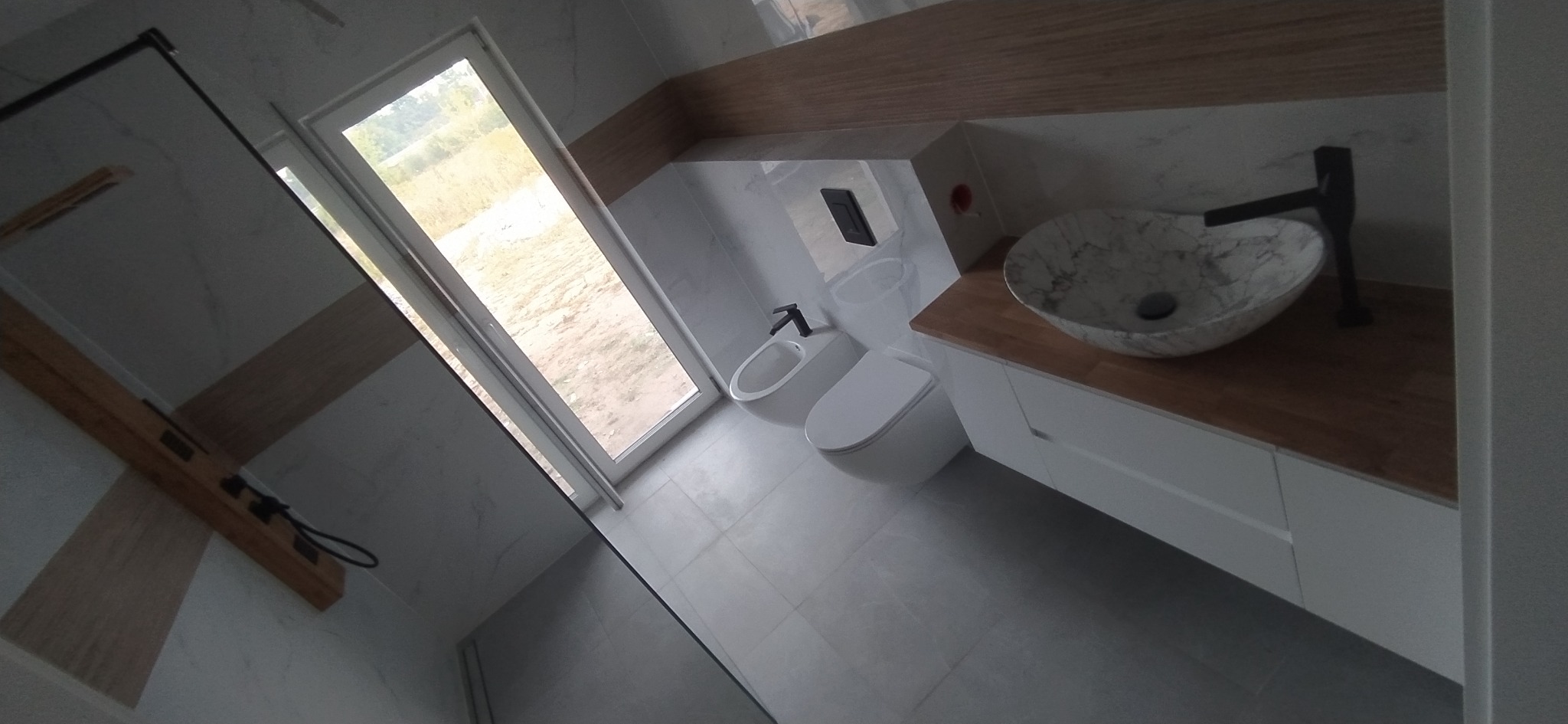 Nowoczesna łazienka z marmurową umywalką, czarną armaturą, białymi meblami i dużym oknem z widokiem na zewnątrz.