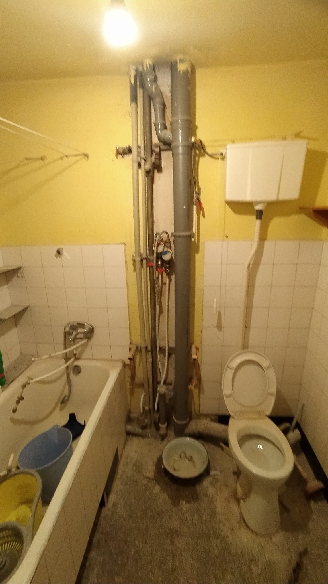 Łazienka w trakcie prac remontowych: widoczne rury, instalacje, wanna z wiadrami, toaleta z otwartą klapą i ślady po kuciu płytek na ścianach.
