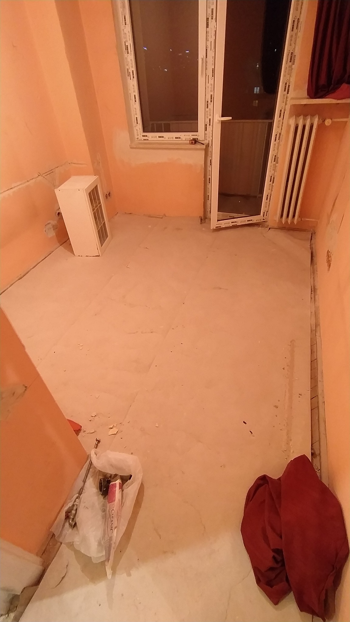 Pokój w trakcie prac przygotowawczych do remontu, widoczne ściany w kolorze brzoskwiniowym, częściowo przygotowane pod malowanie, na podłodze płyty gipsowe, okno balkonowe z widokiem na nocne...