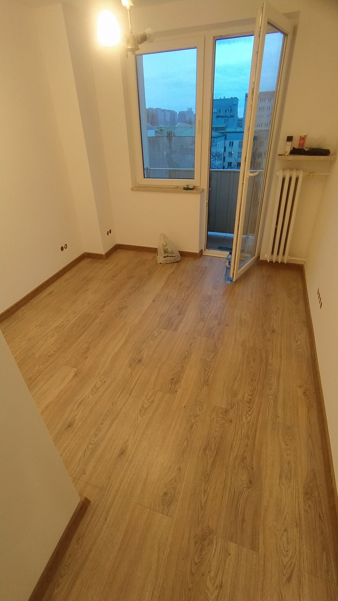Pusty pokój po remoncie z panelami podłogowymi w odcieniach dębu, widok na balkon i miasto w tle przez otwarte drzwi balkonowe, grzejnik na ścianie, widoczne puszki elektryczne.