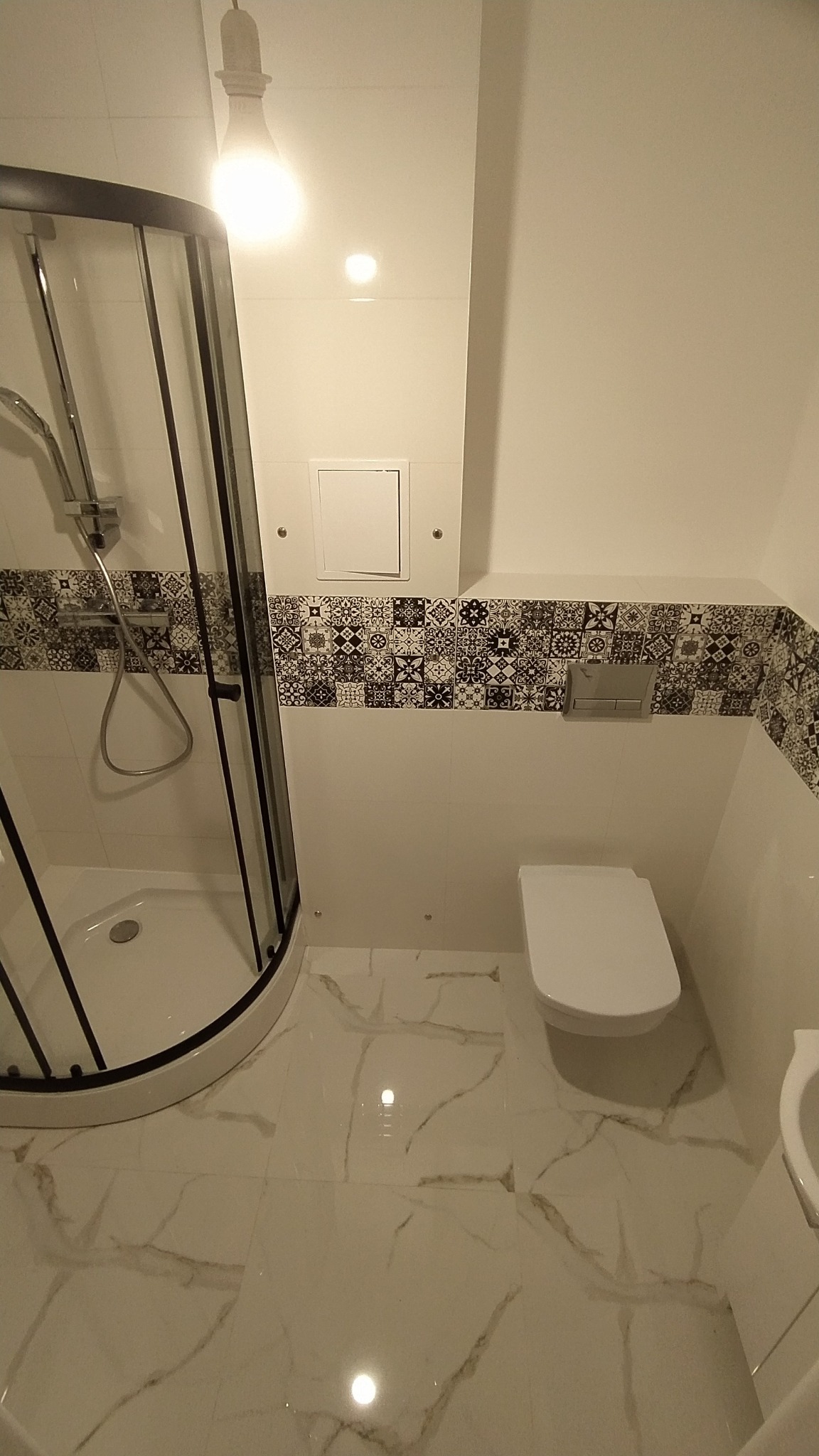 Nowoczesna łazienka z kabiną prysznicową z czarnymi ramami, podwieszaną toaletą i wzorzystymi płytkami dekoracyjnymi w pasie wzdłuż ściany, oświetlona pojedynczą żarówką.