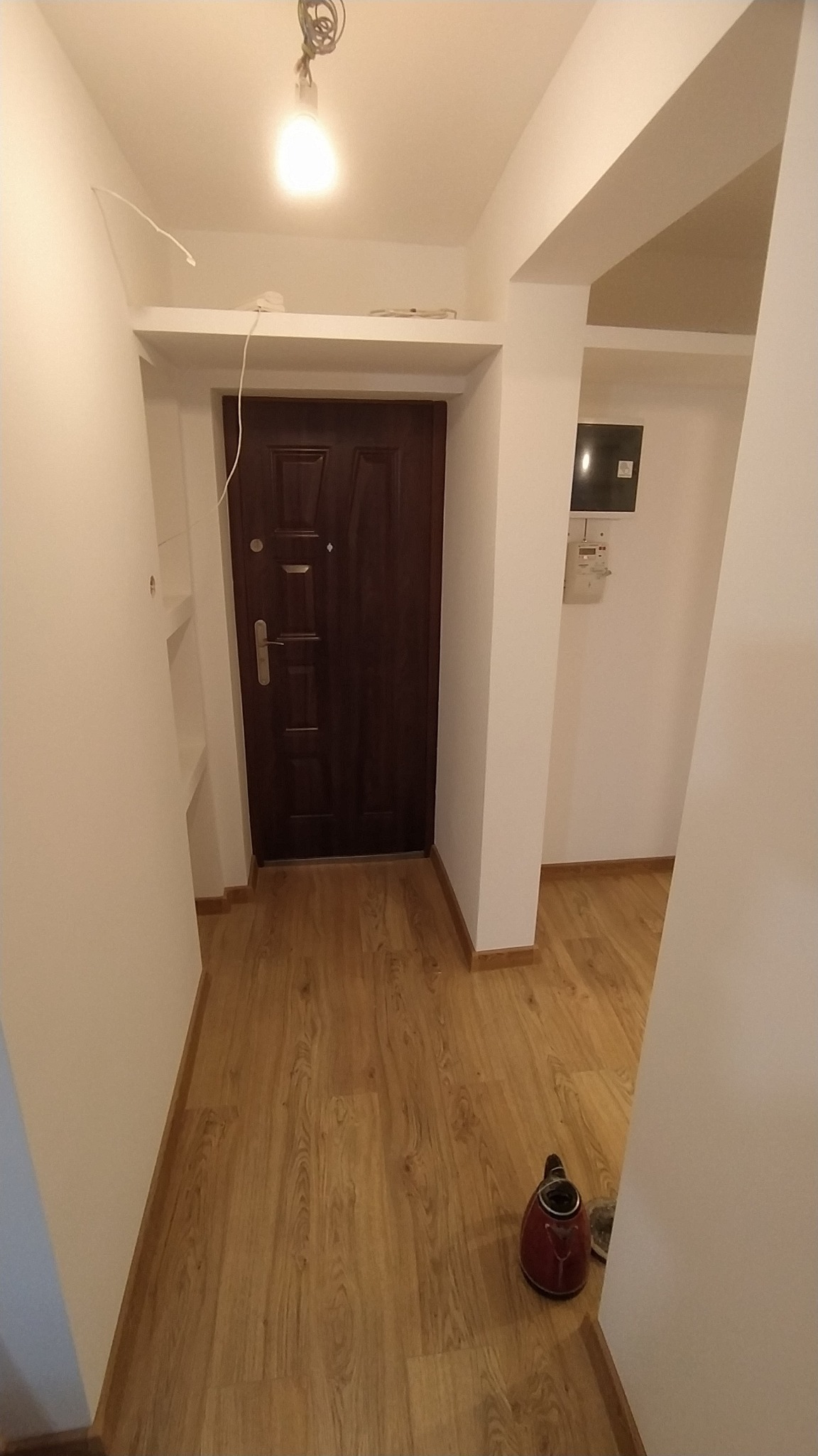 Korytarz po odświeżeniu z drewnopodobną podłogą, białe ściany, ciemne drzwi wejściowe z metalową ościeżnicą, widoczny także fragment instalacji elektrycznej pod sufitem i czerwony czajnik...