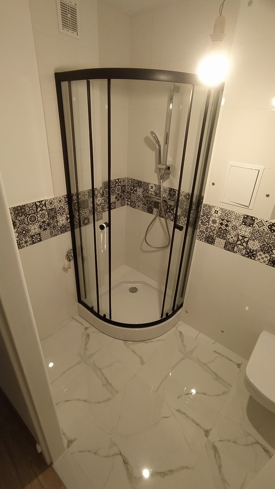 Nowoczesna łazienka z narożną kabiną prysznicową z czarnymi profilami, dekoracyjnym pasem płytek z motywem etnicznym i błyszczącymi marmurkowymi płytkami podłogowymi.