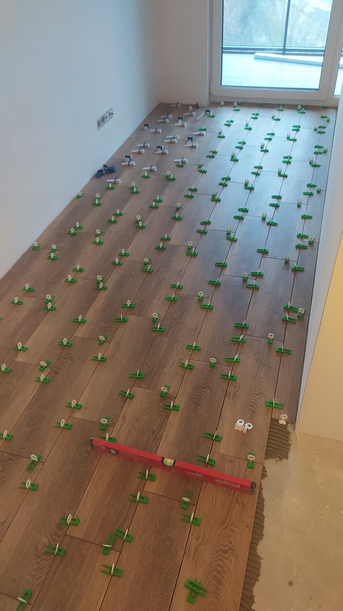 Układanie brązowych płytek podłogowych imitujących drewno z użyciem systemu poziomowania płytek (klipsy i kliny), widoczna poziomica w kolorze czerwonym.