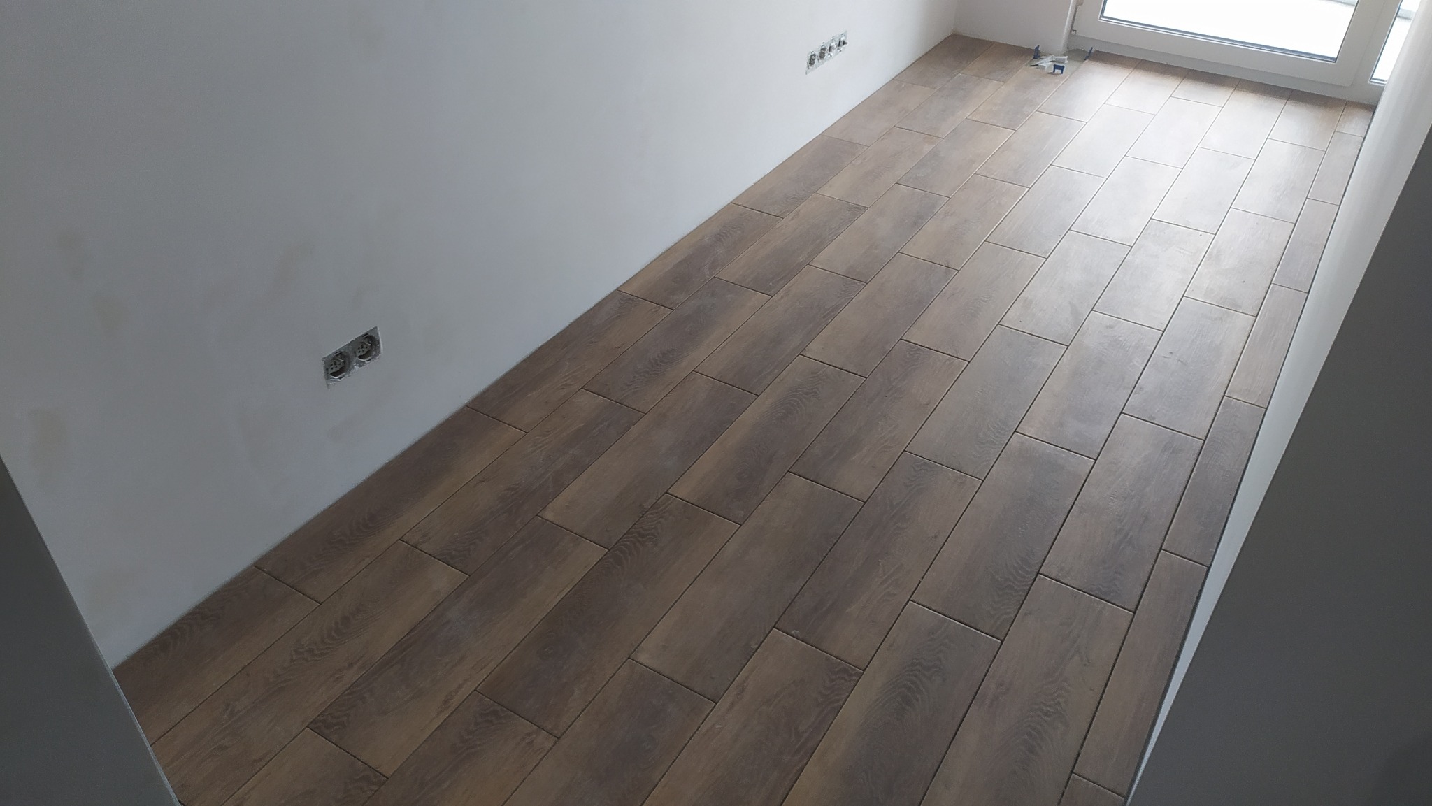 Ułożona podłoga z płytek imitujących drewno w nowym pomieszczeniu, widoczne gniazdka elektryczne na ścianie i fragment okna.