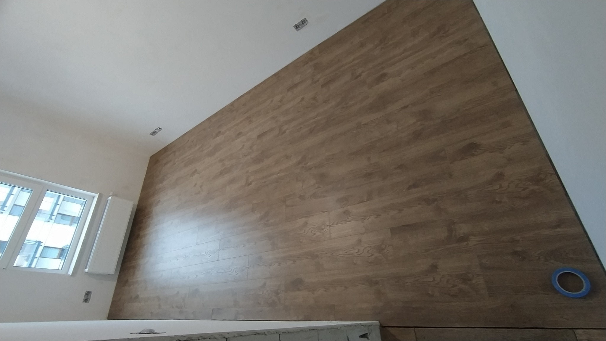 Ściana wykończona panelami imitującymi drewno w jasnym odcieniu, widoczne punkty oświetleniowe na suficie i okno z widokiem na budynki.