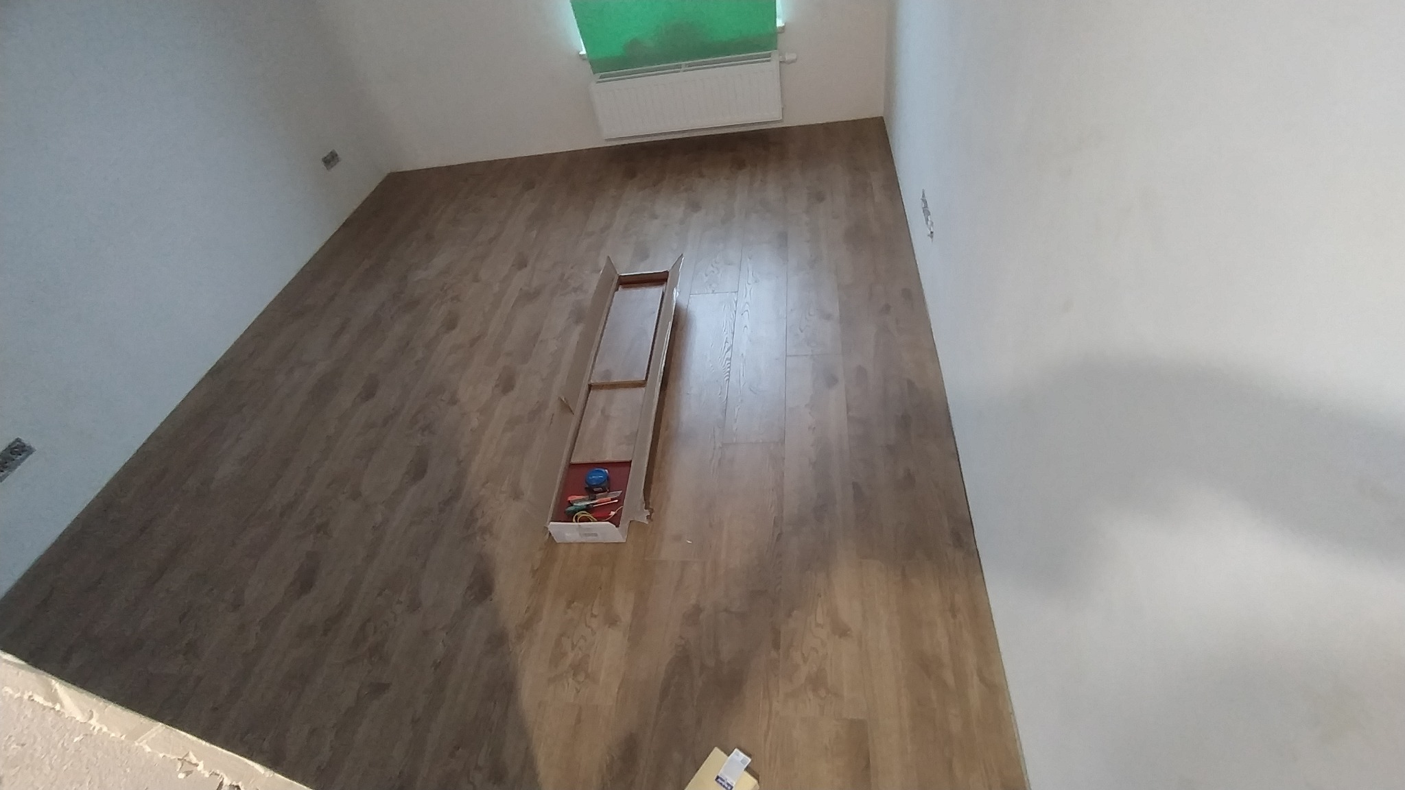 Układanie paneli podłogowych w pokoju, widoczny fragment ułożonej podłogi, otwarte opakowanie z panelami i narzędziami do montażu przy grzejniku.