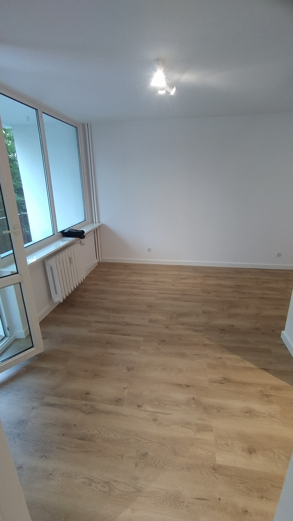 Jasne panele podłogowe z wyraźnym wzorem drewna w pustym, świeżo wykończonym pokoju z białymi ścianami, grzejnikiem pod oknem i nowoczesnym oświetleniem sufitowym.