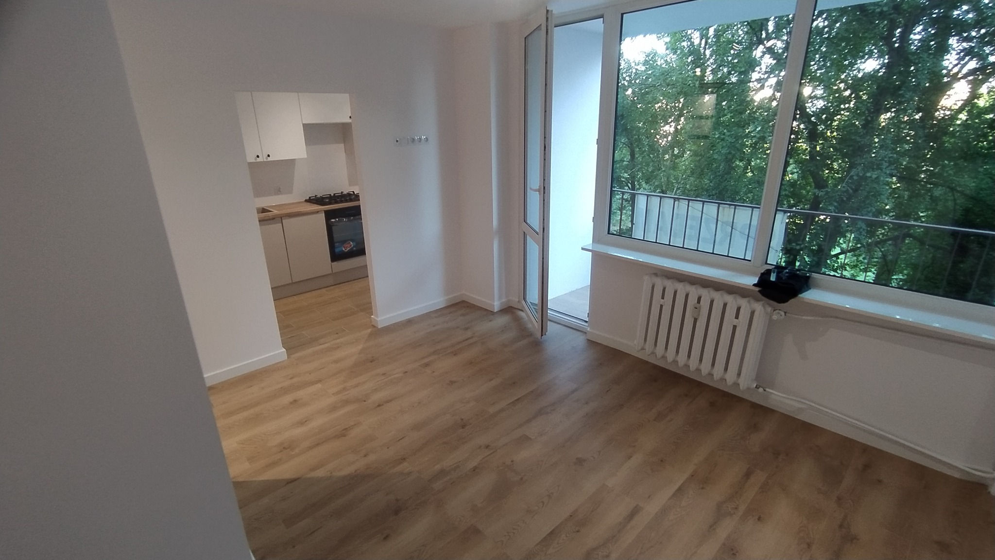 Jasne, odnowione mieszkanie z drewnianą podłogą, widokiem na balkon i zielone drzewa za oknem, oraz białą kuchnią w tle.