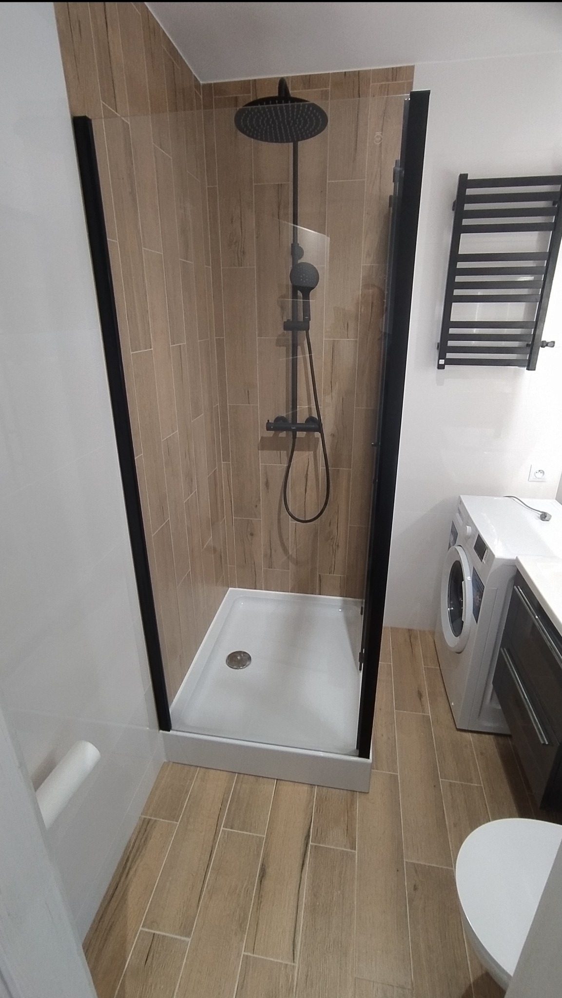 Nowoczesna kabina prysznicowa z czarnymi profilami i deszczownicą, drewnopodobne płytki na ścianie, grzejnik drabinkowy i pralka w tle.