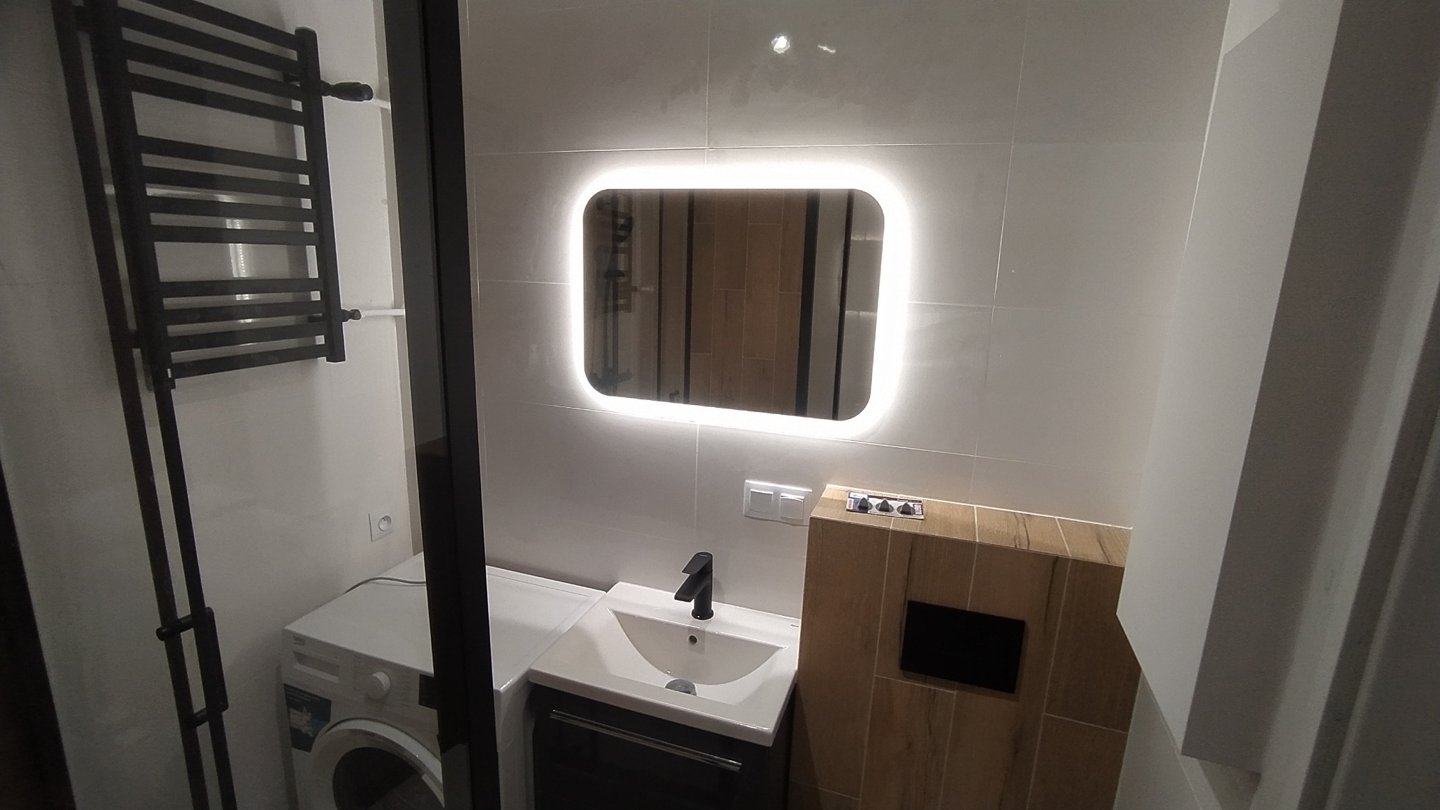 Nowoczesna łazienka z podświetlanym lustrem, umywalką z czarną armaturą, grzejnikiem drabinkowym i pralką w zabudowie.
