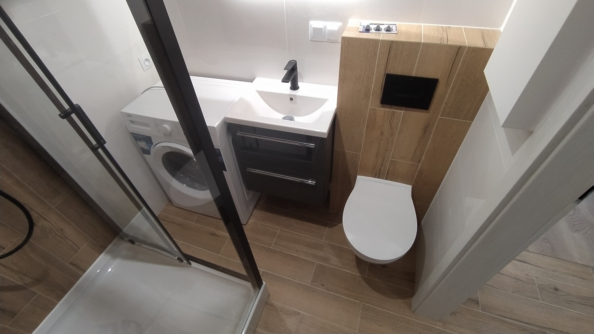 Nowoczesna łazienka z kabiną prysznicową z czarnymi profilami, pralką, umywalką z szafką w kolorze grafitowym i podwieszaną toaletą, wykończona płytkami imitującymi drewno.